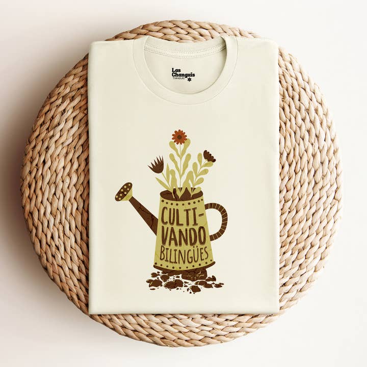 A folded "Cultivando Bilingües" T-Shirt displayed neatly on a flat surface. The shirt features a charming design of a watering pot with the phrase "Cultivando Bilingües" surrounded by blossoming flowers. The fabric appears soft and high-quality, emphasizing its comfort.