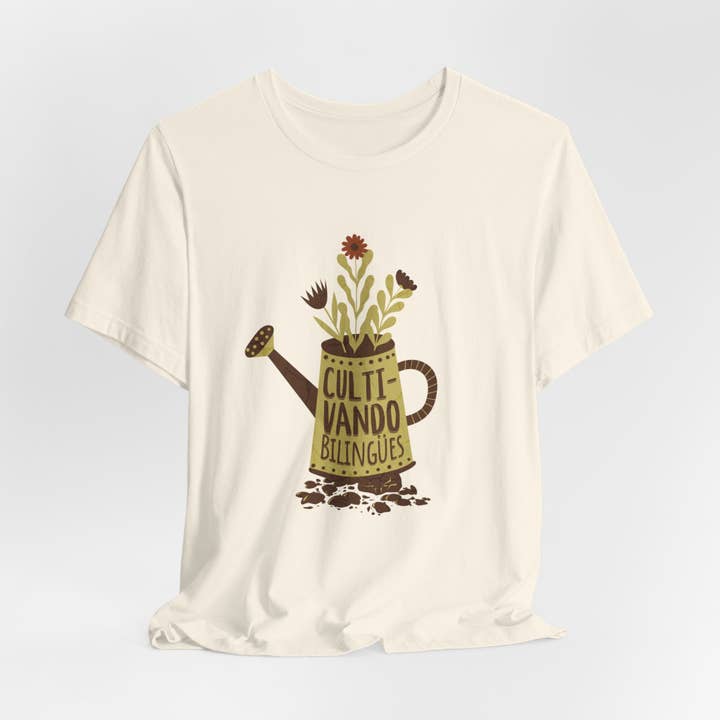 A spread-out "Cultivando Bilingües" T-Shirt laid flat on a textured background. The vibrant design prominently showcases a watering pot with the phrase "Cultivando Bilingües" and colorful flowers, symbolizing the nurturing of growth and language learning. The soft, high-quality cotton fabric is visible, highlighting its durability and comfort.