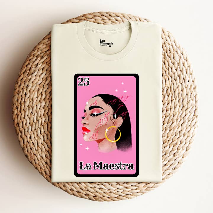 A folded La Maestra Lotería-Inspired T-Shirt displayed on a textured rope mat. The shirt features a colorful design showcasing the number 25 and an illustration of a Latina woman representing "La Maestra." The fabric appears soft and high-quality, emphasizing its comfort.