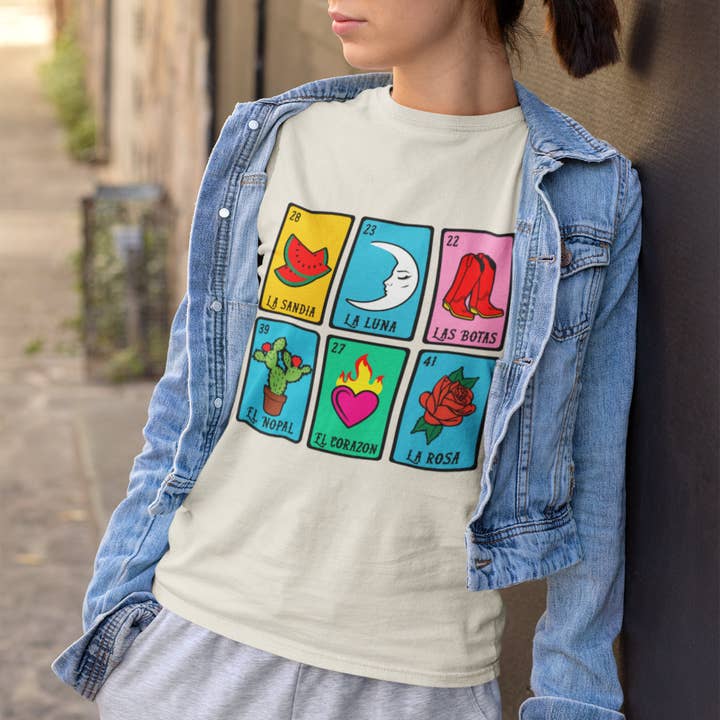 A model stands wearing a colorful Mexican Lotería t-shirt, smiling and showcasing the vibrant designs. The shirt is paired with casual denim shorts, emphasizing a playful and comfortable style, perfect for everyday wear. The background is a lively outdoor setting, enhancing the festive vibe of the outfit.