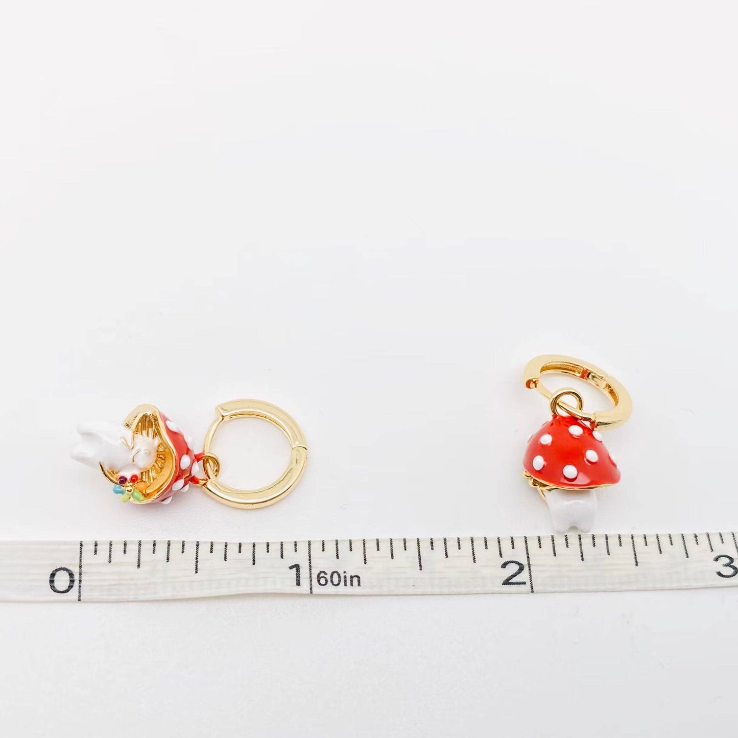 Gold-plated whimsical mushroom enamel earrings in vibrant colors, featuring playful and nature-inspired details, perfect for adding a fun pop of color to any outfit. Earrings showcased next to a measuring tape.
