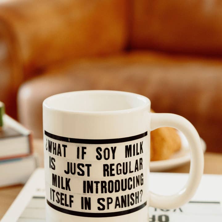 A ceramic mug with the text "Soy Milk" in a simple, modern font. The mug is white with a clean design, perfect for enjoying plant-based beverages. Shown in a cozy setting with a blurry couch in the background.