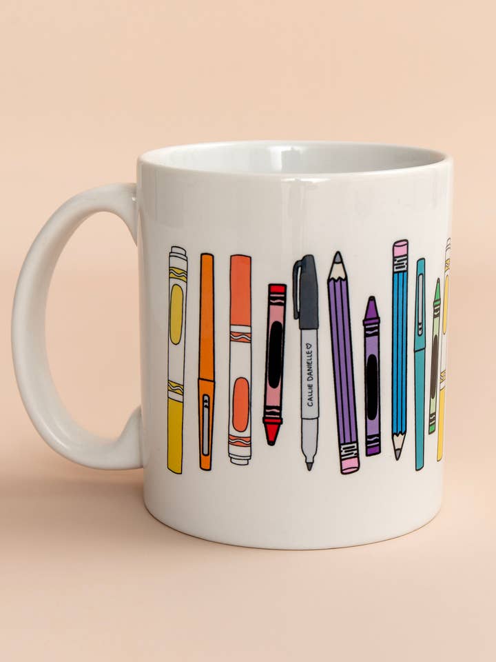 A white mug decorated with colorful illustrations of various writing utensils, including pens and pencils, against a soft background.