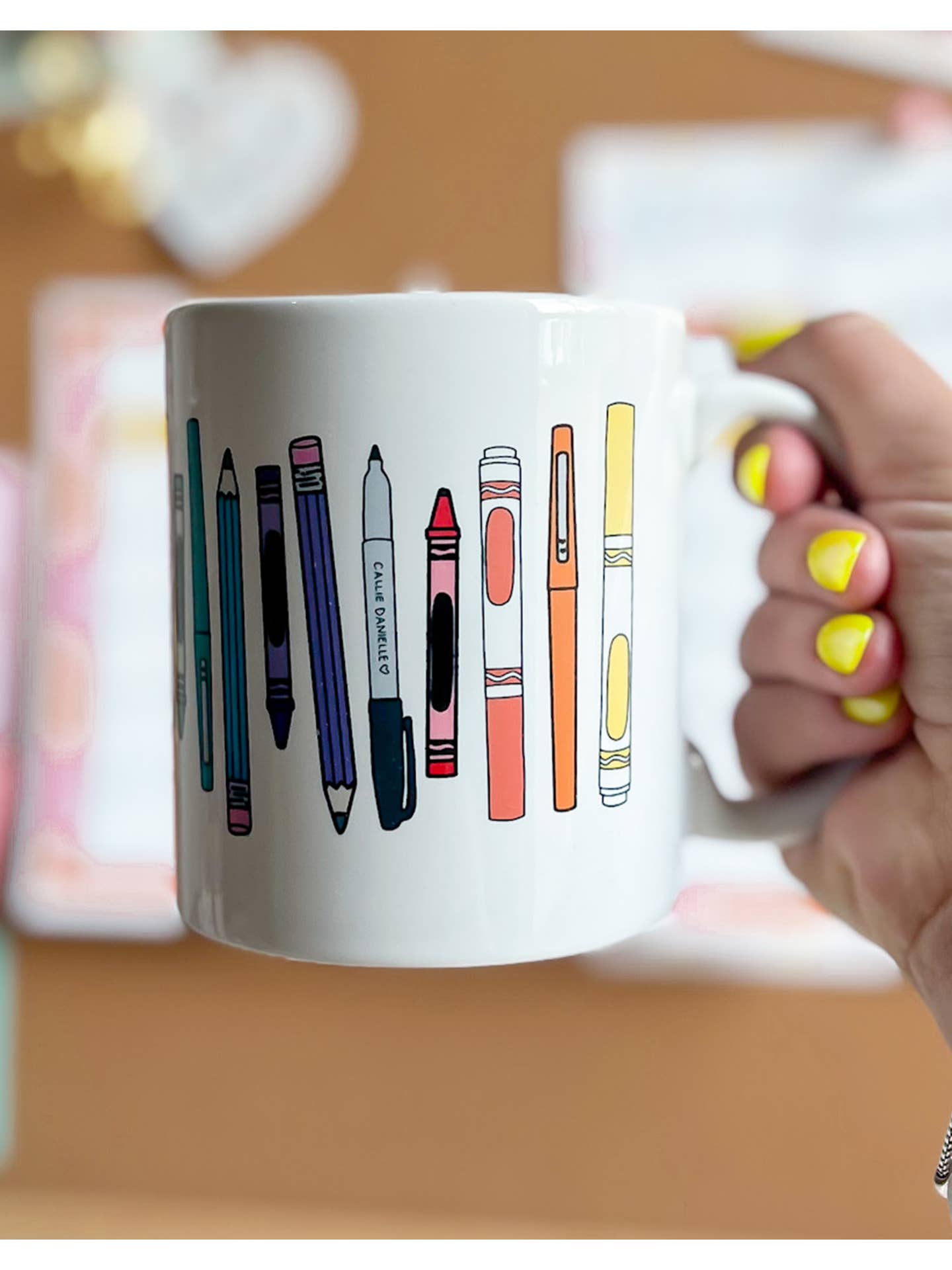 A white mug decorated with colorful illustrations of various writing utensils, including pens and pencils, against a soft background being held by a lady with yellow nails.
