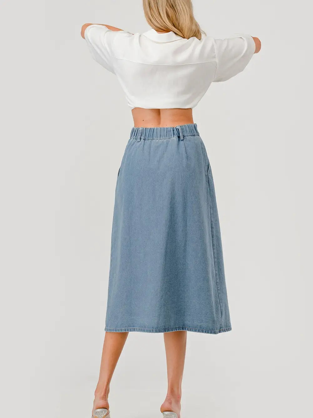 Back view of Woman wearing a white button-up shirt with short sleeves and front pockets, paired with a high-waisted, pleated denim midi skirt.