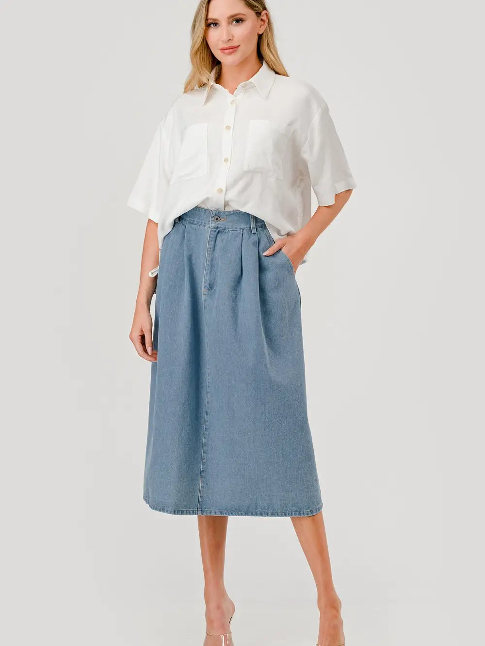 Woman wearing a white button-up shirt with short sleeves and front pockets, paired with a high-waisted, pleated denim midi skirt.