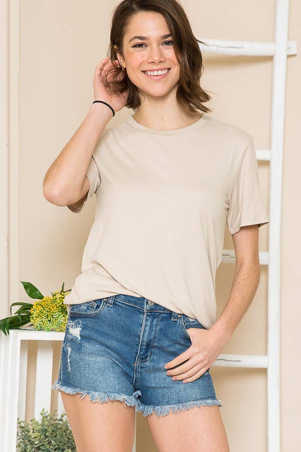 Woman smiling and wearing a beige short-sleeve top with denim shorts, standing casually.