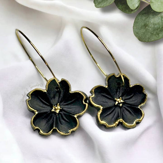 A pair of black floral hoop earrings with gold accents, displayed on a white fabric background.