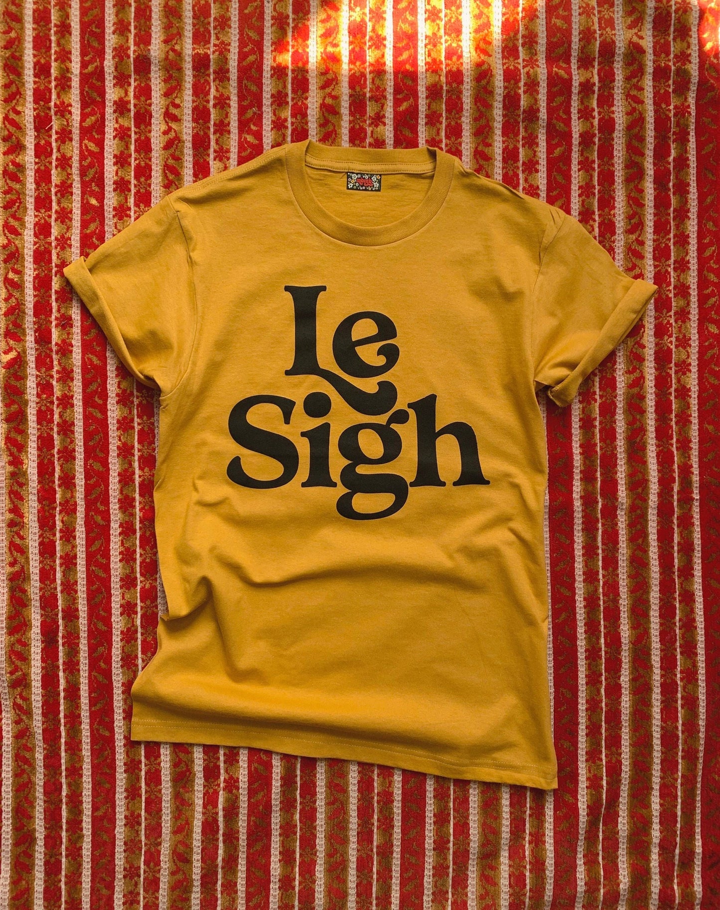  Full view of a mustard yellow "Le Sigh" t-shirt with bold black lettering, neatly laid out on a patterned red and white cloth background.