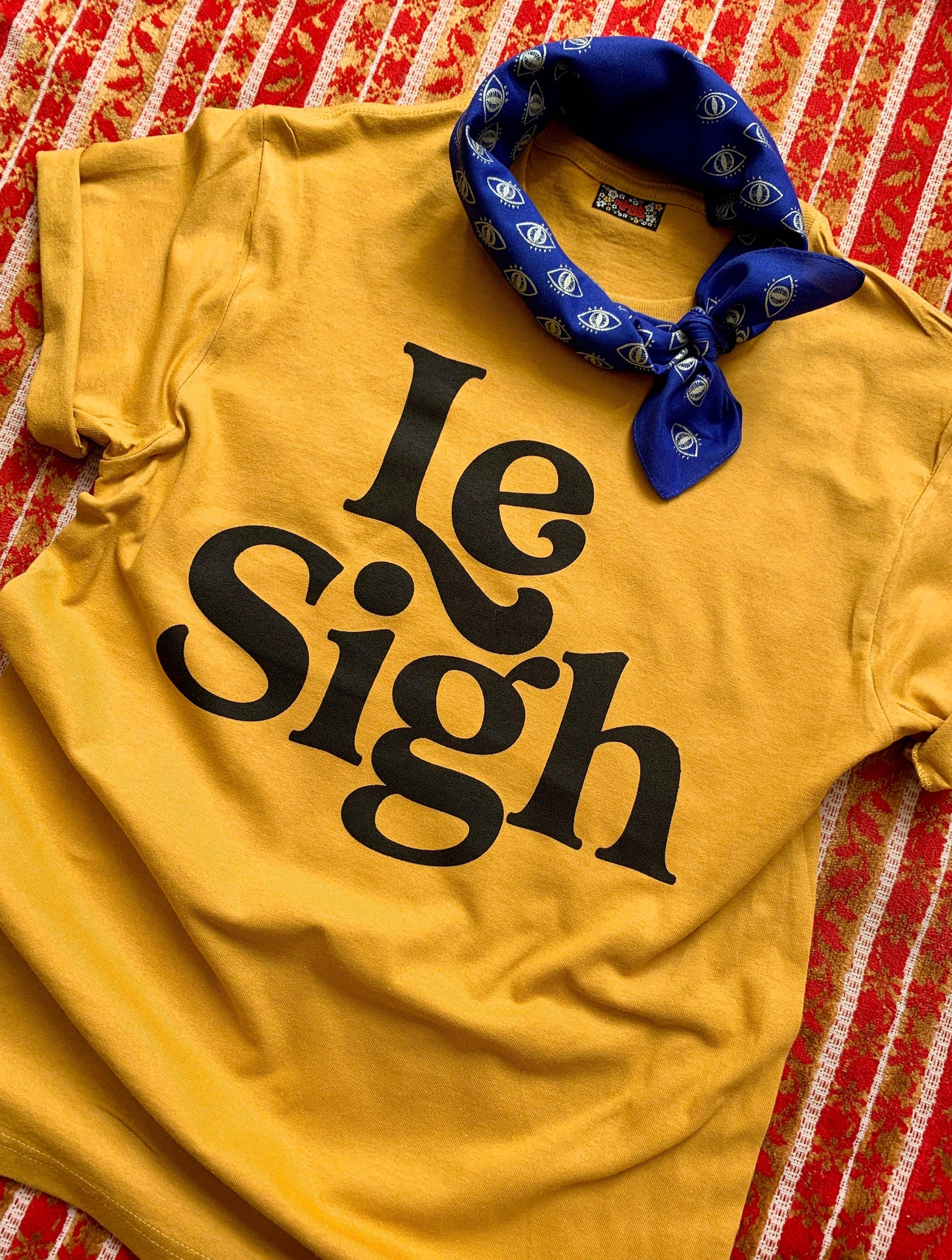 Mustard yellow "Le Sigh" t-shirt styled with a blue bandana, displayed on a patterned red and white cloth background.