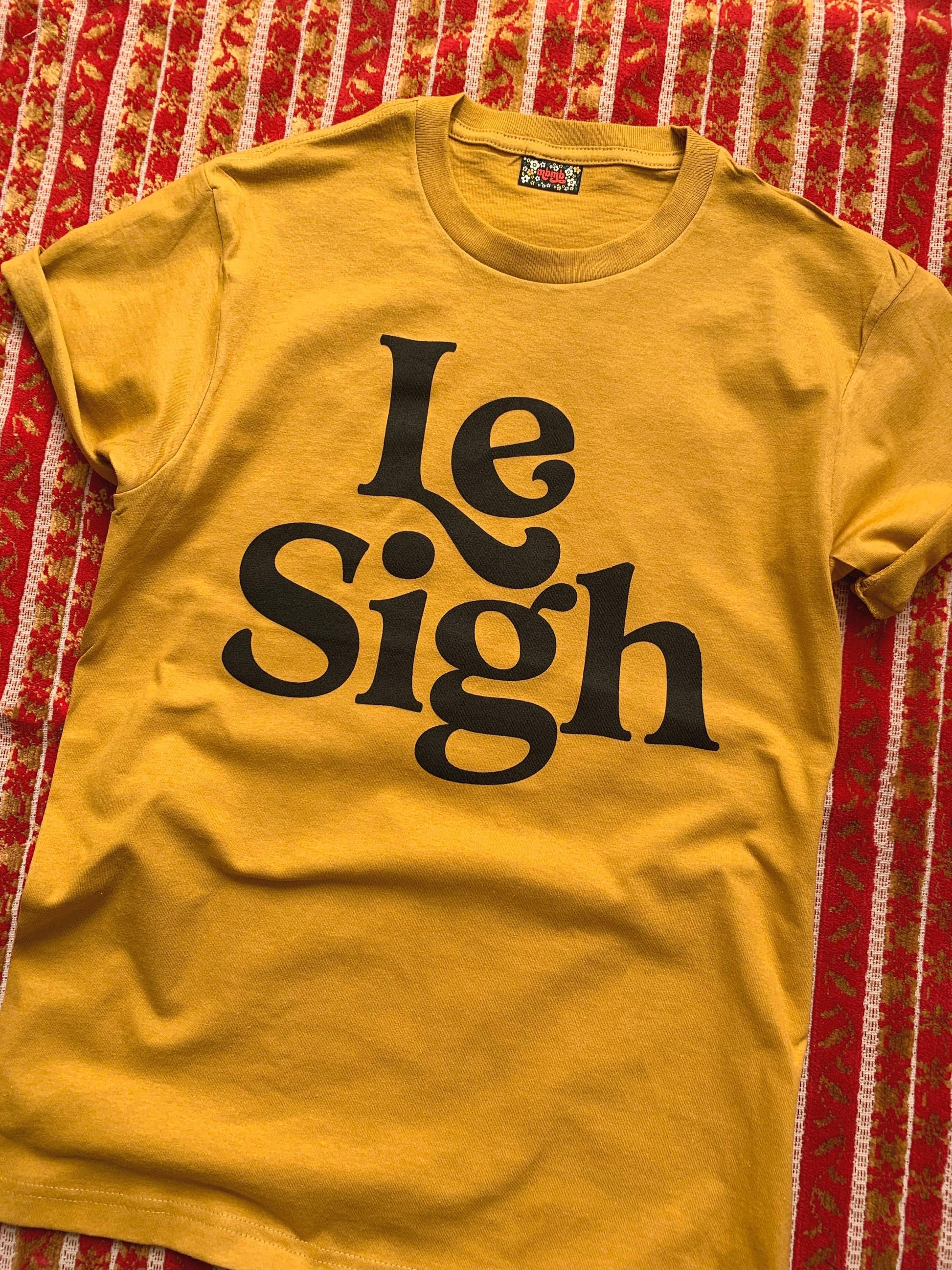 Close-up of a mustard yellow "Le Sigh" t-shirt with bold black lettering, laid flat on a patterned red and white cloth background.