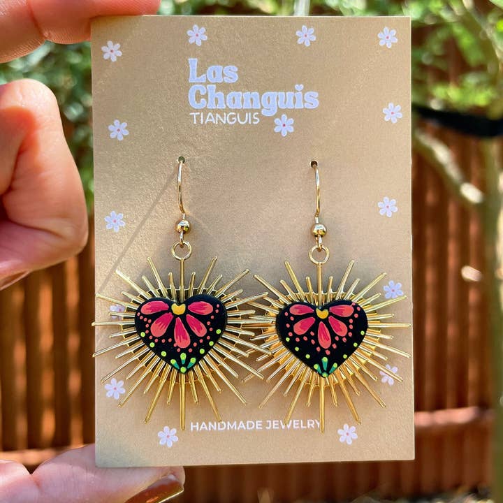 A pair of handmade Sacred Heart earrings crafted from polymer clay, featuring vibrant hand-painted floral details inspired by Mexican folk art. Gold-plated metal spikes radiate from the center, evoking the Sacred Heart symbol, adding a touch of cultural significance and artisan craftsmanship to these stunning earrings.