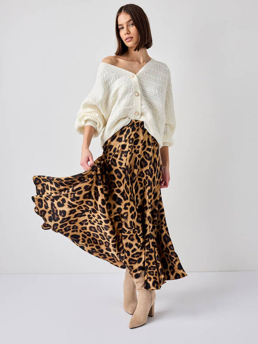 A model twirling in a brown leopard print satin midi skirt, styled with a white off-the-shoulder knit sweater and beige heeled ankle boots. The motion captures the skirt's flowy movement, adding a dynamic element to the chic ensemble.