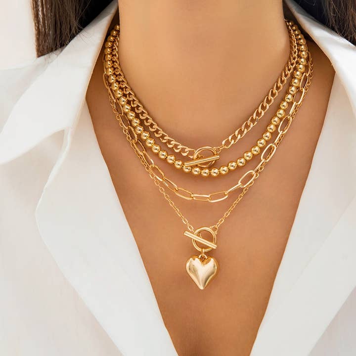 A model wearing a layered gold necklace set with geometric chains and an OT buckle heart pendant, styled with a white blouse.