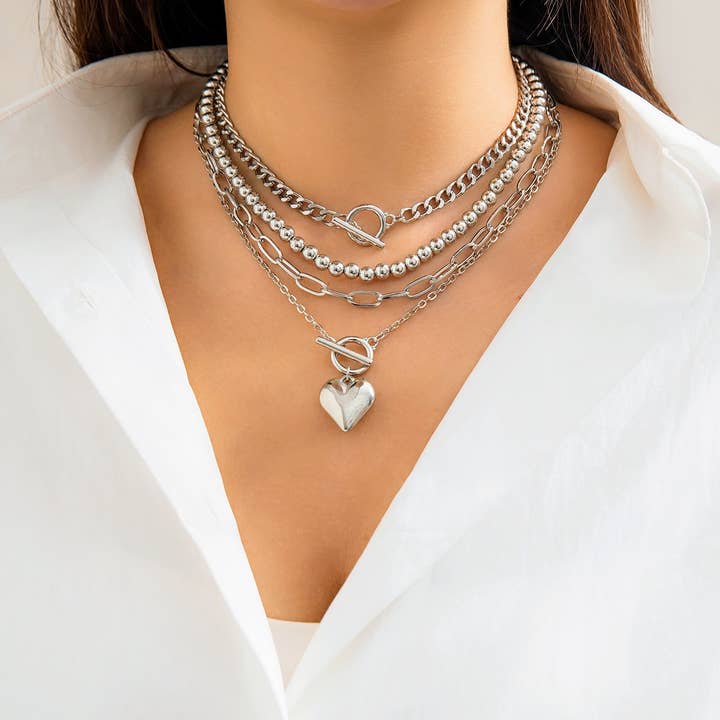 A model showcasing a layered silver necklace set with geometric chains and an OT buckle heart pendant, paired with a white blouse.