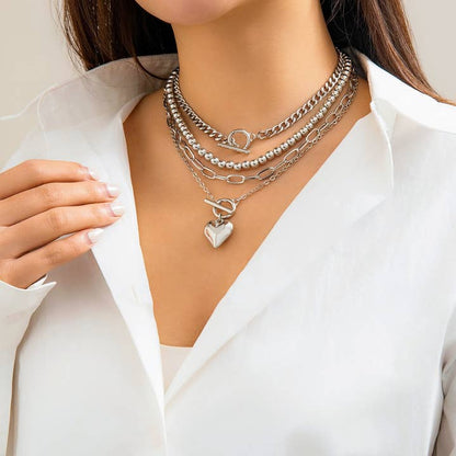 Close-up of a model wearing a layered silver necklace set with geometric chains and an OT buckle heart pendant.