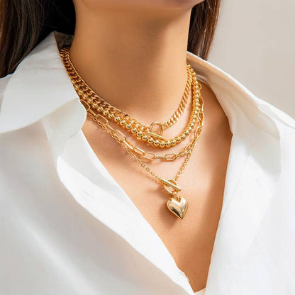 A model displaying a layered gold necklace set with geometric chains and an OT buckle heart pendant, styled with a white blouse.