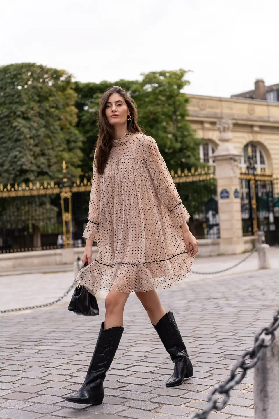 Model wearing a short Iris dress in polka dot tulle featuring black and beige two-tone fabric, three-quarter sleeves, and a flared cut. The dress includes a removable velvet belt at the waist and an inner lining, offering an elegant and sophisticated look with a playful touch of transparency paired with western style boots and black clutch.