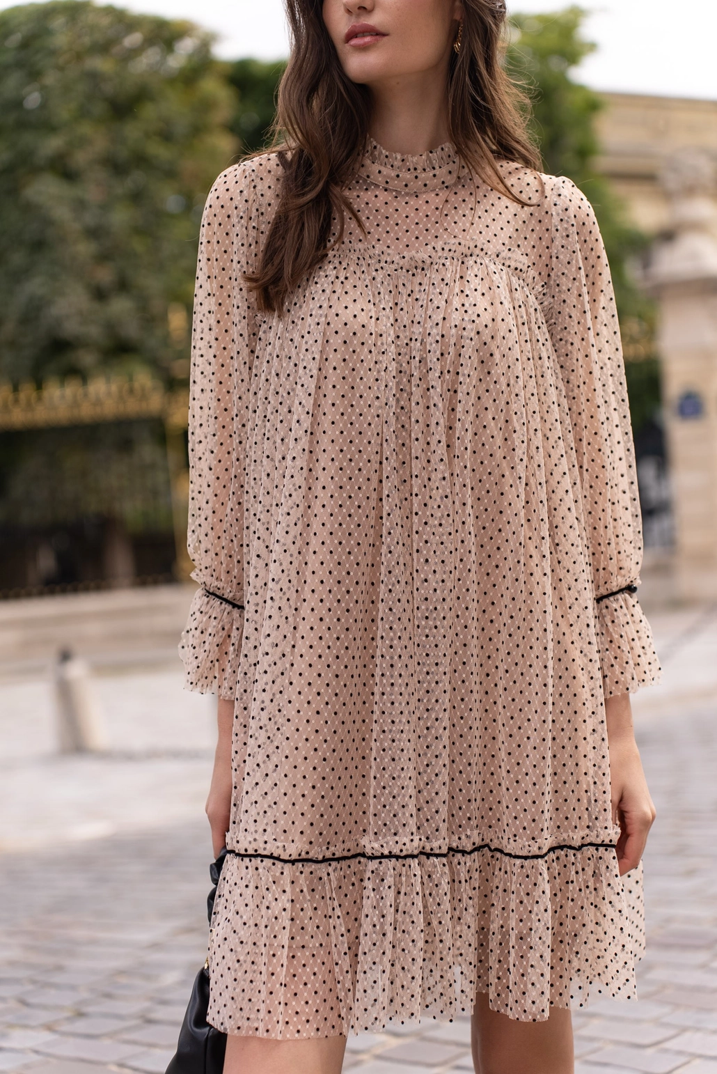 Close up of model wearing a short Iris dress in polka dot tulle featuring black and beige two-tone fabric, three-quarter sleeves, and a flared cut. The dress includes a removable velvet belt at the waist and an inner lining, offering an elegant and sophisticated look with a playful touch of transparency paired with western style boots and black clutch.